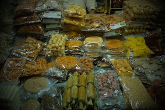 The image displays an assortment of packaged snacks and sweets, including various types of chips, cookies, and other savory and sweet items, neatly stacked and organized in plastic bags. They are placed on a table likely in a market or store setting.