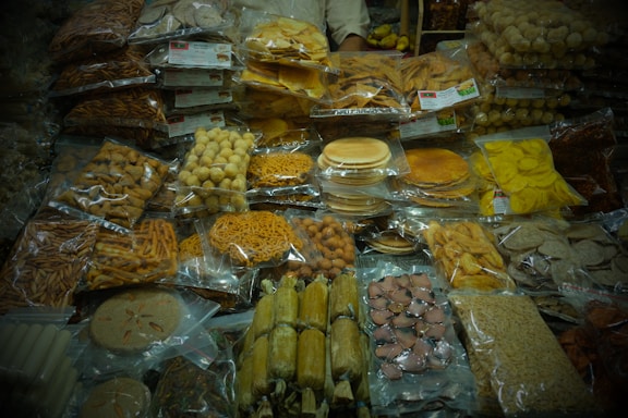The image displays an assortment of packaged snacks and sweets, including various types of chips, cookies, and other savory and sweet items, neatly stacked and organized in plastic bags. They are placed on a table likely in a market or store setting.