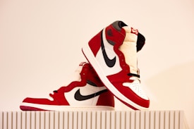 A pair of red, white, and black high-top sneakers are displayed on a ribbed white surface. The design features a prominent black swoosh logo on the side and red accents with a white base. They are stylishly positioned, one leaning against the other.
