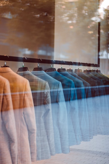 A row of suits hanging on a rack inside a store, viewed through a glass window. The reflection on the glass creates a blend of indoor and outdoor elements, adding an artistic overlay to the display.