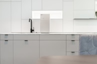 A contemporary kitchen interior featuring minimalist design with clean lines. The cabinets are sleek and matte in finish, predominantly white with a hint of gray. A black faucet is installed on the countertop, which is made of a light-colored marble material. A modern water filter unit is mounted on the wall above the counter. The overall lighting is bright, providing a spacious and airy feel.