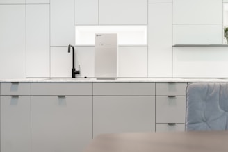 A contemporary kitchen interior featuring minimalist design with clean lines. The cabinets are sleek and matte in finish, predominantly white with a hint of gray. A black faucet is installed on the countertop, which is made of a light-colored marble material. A modern water filter unit is mounted on the wall above the counter. The overall lighting is bright, providing a spacious and airy feel.