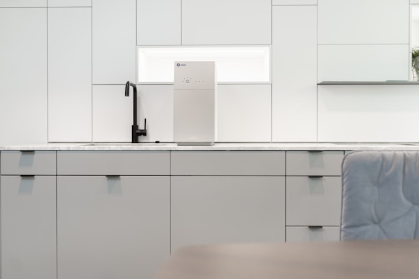 A contemporary kitchen interior featuring minimalist design with clean lines. The cabinets are sleek and matte in finish, predominantly white with a hint of gray. A black faucet is installed on the countertop, which is made of a light-colored marble material. A modern water filter unit is mounted on the wall above the counter. The overall lighting is bright, providing a spacious and airy feel.