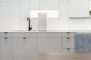 A contemporary kitchen interior featuring minimalist design with clean lines. The cabinets are sleek and matte in finish, predominantly white with a hint of gray. A black faucet is installed on the countertop, which is made of a light-colored marble material. A modern water filter unit is mounted on the wall above the counter. The overall lighting is bright, providing a spacious and airy feel.