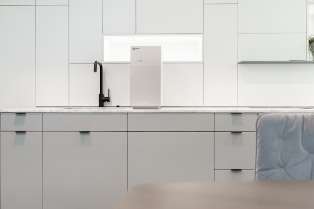 A contemporary kitchen interior featuring minimalist design with clean lines. The cabinets are sleek and matte in finish, predominantly white with a hint of gray. A black faucet is installed on the countertop, which is made of a light-colored marble material. A modern water filter unit is mounted on the wall above the counter. The overall lighting is bright, providing a spacious and airy feel.