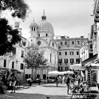 A black and white photograph captures a charming European square with a central domed church and surrounding historic buildings. People casually stroll or gather in the area, and outdoor seating is visible under umbrellas. Trees provide some greenery amidst the stone architecture.