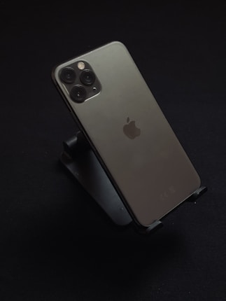 An iPhone with a sleek design featuring three camera lenses on the back, resting on a dark stand against a black background. The Apple logo is prominently displayed.