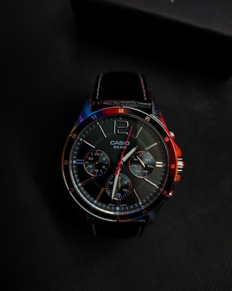 A close-up view of a Casio wristwatch with a black leather strap. The dial features multiple sub-dials with various markings for timekeeping functionalities. The watch is illuminated with colorful reflections of blue, red, and orange light.