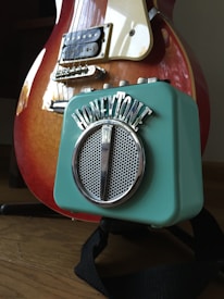 A close-up of an electric guitar and a small turquoise amplifier. The guitar has a sunburst finish with a white pickguard and metal strings. The amplifier has a vintage design with the brand name 'HONEYTONE' displayed prominently on the top, and a circular silver speaker grill.