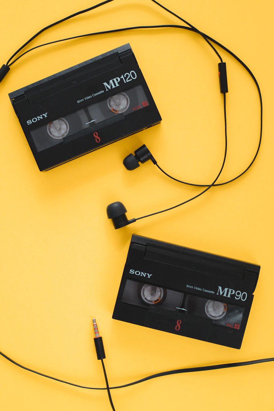 The image presents two black Sony Mp70 headphones, each connected to a separate cassette tape by wires. The tapes are neatly arranged on top of a yellow background, creating an interesting contrast between the vibrant color and the monochrome audio equipment.
