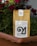 A brown paper coffee bag labeled 'Türk Medium' is placed inside a brown planter. Behind the bag, vivid red flowers and green foliage create a colorful background. There is a logo and text printed in black on the coffee bag.