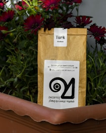 A brown paper coffee bag labeled 'Türk Medium' is placed inside a brown planter. Behind the bag, vivid red flowers and green foliage create a colorful background. There is a logo and text printed in black on the coffee bag.
