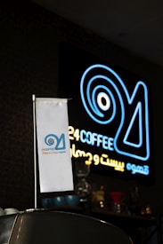 A neon sign with the text '24 COFFEE' glows in blue and yellow colors against a dark background. In the foreground, a small white flag with a logo and the same text is positioned on a counter, along with various coffee-related equipment.