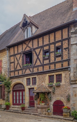 A traditional timber-framed building with a steep, tiled roof and multiple windows adorned with decorative shutters. The facade features exposed wooden beams crisscrossing over a brick and plaster wall. There are potted plants and flowers placed around the entrance, adding a touch of greenery. A small stone well with a shingled roof is positioned near the entrance, enhancing the rustic charm.