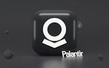 A dark, glossy cube with a white abstract symbol on its front surface, resembling a stylized human figure. The word 'Palantir' is written in white at the base of the cube in a three-dimensional font. Surrounding the cube are smaller, floating spheres that add depth to the composition.