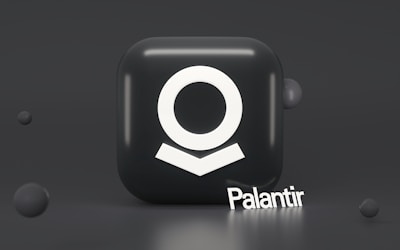 A dark, glossy cube with a white abstract symbol on its front surface, resembling a stylized human figure. The word 'Palantir' is written in white at the base of the cube in a three-dimensional font. Surrounding the cube are smaller, floating spheres that add depth to the composition.