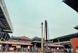 A traditional temple with ornate architecture and a tiled roof is surrounded by people. Two tall stone pillars with intricate carvings are prominently displayed in the foreground. The atmosphere appears busy with several visitors dressed in casual attire moving around near the temple.