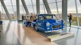 A classic blue Mercedes-Benz truck is displayed indoors, carrying a vintage silver race car. The setting appears to be a modern showroom or museum with large floor-to-ceiling windows, providing a view of the cityscape outside. Two individuals stand near the vehicles, one photographing the scene.