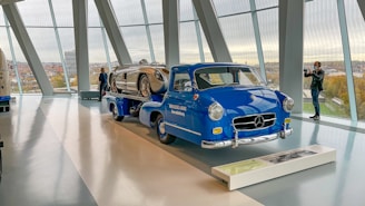 A classic blue Mercedes-Benz truck is displayed indoors, carrying a vintage silver race car. The setting appears to be a modern showroom or museum with large floor-to-ceiling windows, providing a view of the cityscape outside. Two individuals stand near the vehicles, one photographing the scene.