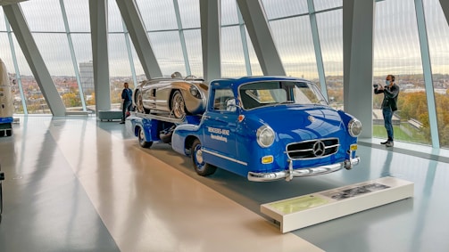 A classic blue Mercedes-Benz truck is displayed indoors, carrying a vintage silver race car. The setting appears to be a modern showroom or museum with large floor-to-ceiling windows, providing a view of the cityscape outside. Two individuals stand near the vehicles, one photographing the scene.