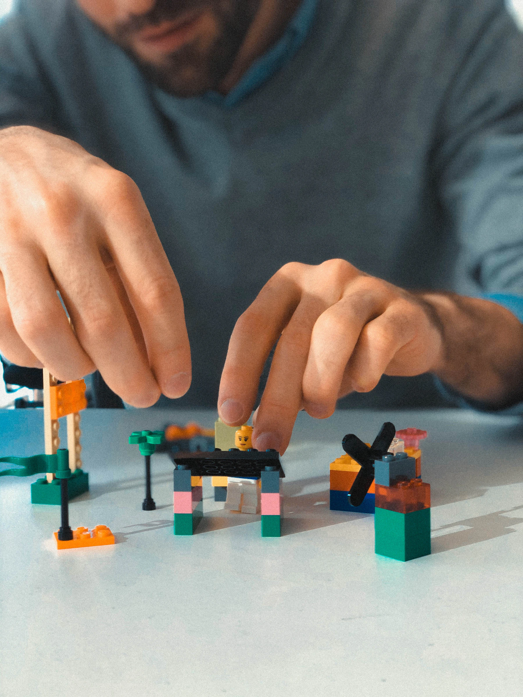 A man in a gray sweater placing a brick on one of three small models on a white table