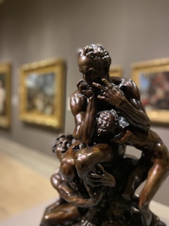 A bronze sculpture depicting a dramatic scene with intertwined figures displaying intense emotion. The intricate details in the musculature and expressions convey movement and struggle, while the gallery setting includes blurred paintings in the background.