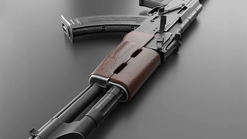 A close-up view of a rifle with a wooden grip and a curved magazine. The weapon is displayed on a smooth, grey surface, showing details of its metallic and wooden parts.