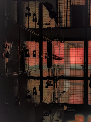 A dimly lit corridor of lockers lines one side, with a series of padlocks securing each locker. The light casts a reddish glow, reflecting on the glossy surface of the metallic doors. The far end reveals a barred gate, enhancing the sense of enclosure.