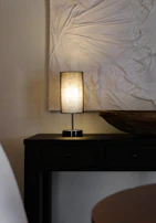 Close-up of a matte black minimalist lamp casting a warm glow on an earthy-toned wooden side table.