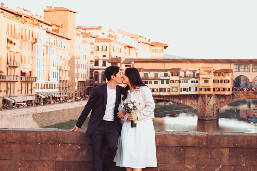 a man and woman kissing on a bridge over a river with buildings