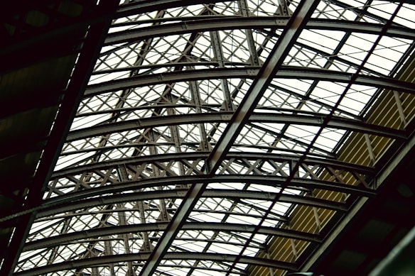 A complex and intricate metal framework forming an overhead structure, featuring interlocking beams and girders. The design conveys a sense of architectural strength and industrial sophistication.