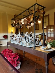 A gingerbread house model is displayed on a wooden table in a traditional kitchen setting. Above the table, an assortment of copper pots and pans hang from a black metal rack. To the left, there are vibrant red poinsettia plants, adding a festive touch. On the right side of the table, there is a vase with red flowers and a plate of cookies next to a bowl of cranberries.