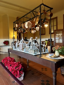 A gingerbread house model is displayed on a wooden table in a traditional kitchen setting. Above the table, an assortment of copper pots and pans hang from a black metal rack. To the left, there are vibrant red poinsettia plants, adding a festive touch. On the right side of the table, there is a vase with red flowers and a plate of cookies next to a bowl of cranberries.