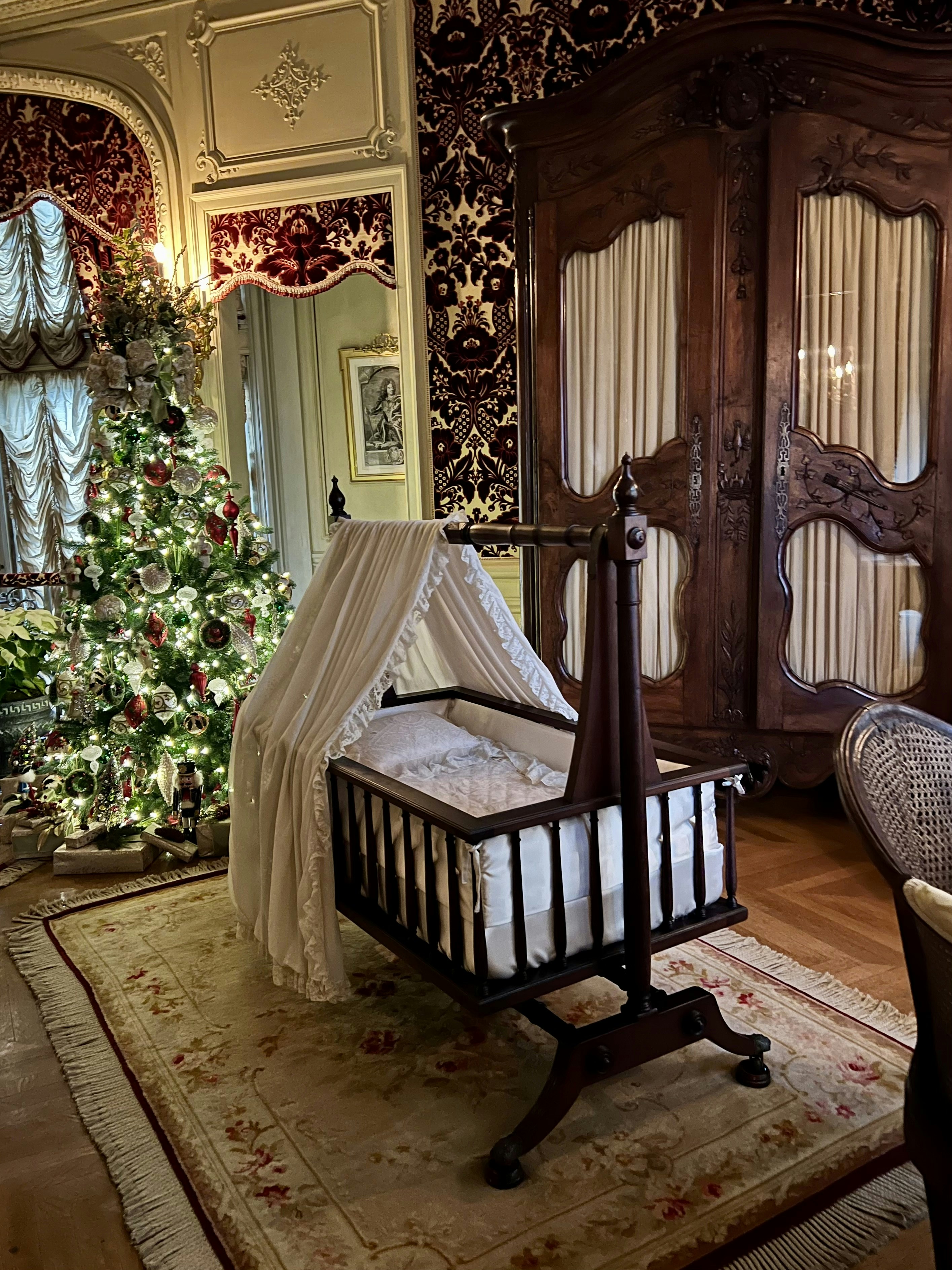 An ornate room featuring a vintage wooden cradle with an elegant canopy. A decorated Christmas tree with sparkling lights and ornaments stands nearby. The walls are adorned with luxurious wallpaper, and a large wooden armoire adds to the antique ambiance. A plush rug covers the floor, enhancing the classic and cozy feel of the space.