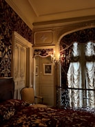 An ornate bedroom with intricate design details, including patterned wallpaper, draped curtains, and an opulent bedspread. The space features an elegant armchair, framed artwork on the walls, and decorative moldings. A vintage chandelier sconce casts a warm glow, enhancing the luxurious ambiance.