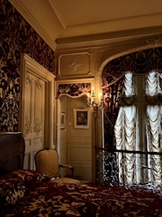 An ornate bedroom with intricate design details, including patterned wallpaper, draped curtains, and an opulent bedspread. The space features an elegant armchair, framed artwork on the walls, and decorative moldings. A vintage chandelier sconce casts a warm glow, enhancing the luxurious ambiance.