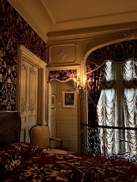 An ornate bedroom with intricate design details, including patterned wallpaper, draped curtains, and an opulent bedspread. The space features an elegant armchair, framed artwork on the walls, and decorative moldings. A vintage chandelier sconce casts a warm glow, enhancing the luxurious ambiance.