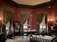 A richly decorated room featuring ornate red wallpaper with intricate patterns, heavy green drapery framing tall windows, and antique wooden furniture. The room includes a small round table adorned with a floral centerpiece, surrounded by elegant chairs. A plush, patterned ottoman sits on a detailed carpet, and a large, ornate mirror stands near some potted plants. Wall sconces provide warm lighting, enhancing the vintage ambiance of the space.