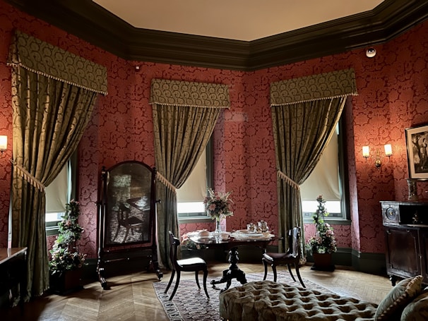 A richly decorated room featuring ornate red wallpaper with intricate patterns, heavy green drapery framing tall windows, and antique wooden furniture. The room includes a small round table adorned with a floral centerpiece, surrounded by elegant chairs. A plush, patterned ottoman sits on a detailed carpet, and a large, ornate mirror stands near some potted plants. Wall sconces provide warm lighting, enhancing the vintage ambiance of the space.