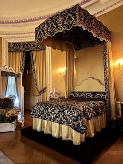 An ornate, luxurious bedroom featuring a lavish, four-poster canopy bed with intricate dark and gold bedding. The room is adorned with elegant drapery and grand decorative elements. A large mirror with an ornate frame rests against the wall beside the bed, adding to the opulent ambiance. A floral arrangement sits on a nearby chair, enhancing the sophisticated decor of the space.