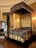 An ornate, luxurious bedroom featuring a lavish, four-poster canopy bed with intricate dark and gold bedding. The room is adorned with elegant drapery and grand decorative elements. A large mirror with an ornate frame rests against the wall beside the bed, adding to the opulent ambiance. A floral arrangement sits on a nearby chair, enhancing the sophisticated decor of the space.