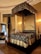 An ornate, luxurious bedroom featuring a lavish, four-poster canopy bed with intricate dark and gold bedding. The room is adorned with elegant drapery and grand decorative elements. A large mirror with an ornate frame rests against the wall beside the bed, adding to the opulent ambiance. A floral arrangement sits on a nearby chair, enhancing the sophisticated decor of the space.