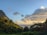 A lush tropical landscape featuring a steep, green mountain on the left and a vibrant sky with scattered clouds. Tall palm trees and dense foliage spread across the foreground, leading to a distant view of the calm ocean under the gentle light of the setting or rising sun.