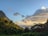 A lush tropical landscape featuring a steep, green mountain on the left and a vibrant sky with scattered clouds. Tall palm trees and dense foliage spread across the foreground, leading to a distant view of the calm ocean under the gentle light of the setting or rising sun.