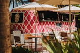 A vibrant pink food truck with a white diamond pattern, labeled 'El Jefe,' is parked under large white umbrellas. Wooden tables and chairs are arranged in front, and lush green foliage is partially visible. The setting appears relaxed and tropical.
