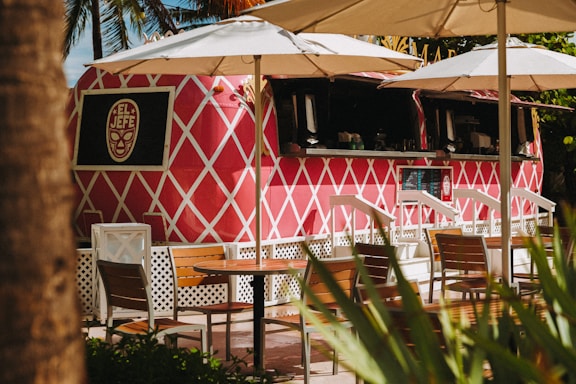 A vibrant pink food truck with a white diamond pattern, labeled 'El Jefe,' is parked under large white umbrellas. Wooden tables and chairs are arranged in front, and lush green foliage is partially visible. The setting appears relaxed and tropical.