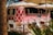 A vibrant pink food truck with a white diamond pattern, labeled 'El Jefe,' is parked under large white umbrellas. Wooden tables and chairs are arranged in front, and lush green foliage is partially visible. The setting appears relaxed and tropical.
