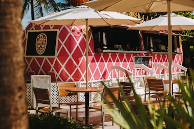 A vibrant pink food truck with a white diamond pattern, labeled 'El Jefe,' is parked under large white umbrellas. Wooden tables and chairs are arranged in front, and lush green foliage is partially visible. The setting appears relaxed and tropical.