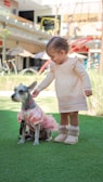 A young child in a cream-colored dress and fluffy boots is gently touching the head of a small dog dressed in a pink outfit. They are standing on a green patch of artificial grass in what appears to be a commercial outdoor area, with blurred backgrounds of benches, umbrellas, and some building structures.