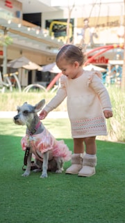 A young child in a cream-colored dress and fluffy boots is gently touching the head of a small dog dressed in a pink outfit. They are standing on a green patch of artificial grass in what appears to be a commercial outdoor area, with blurred backgrounds of benches, umbrellas, and some building structures.
